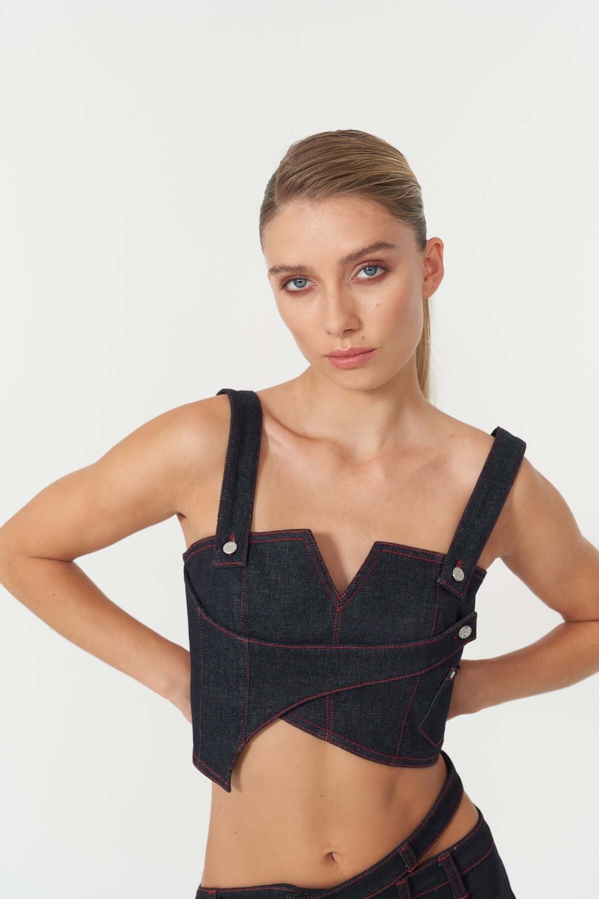 The JENDUE Harvey blue denim crop top showcases a front zipper, wide straps, and decorative silver buttons on the sides.