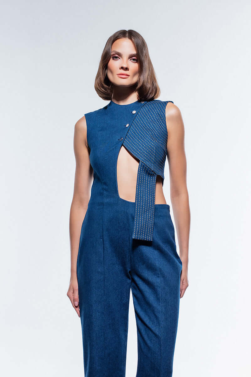 Navy blue denim jumpsuit featuring a sleeveless design and a unique, asymmetric cut-out on the bodice with decorative stitching and silver buttons.
