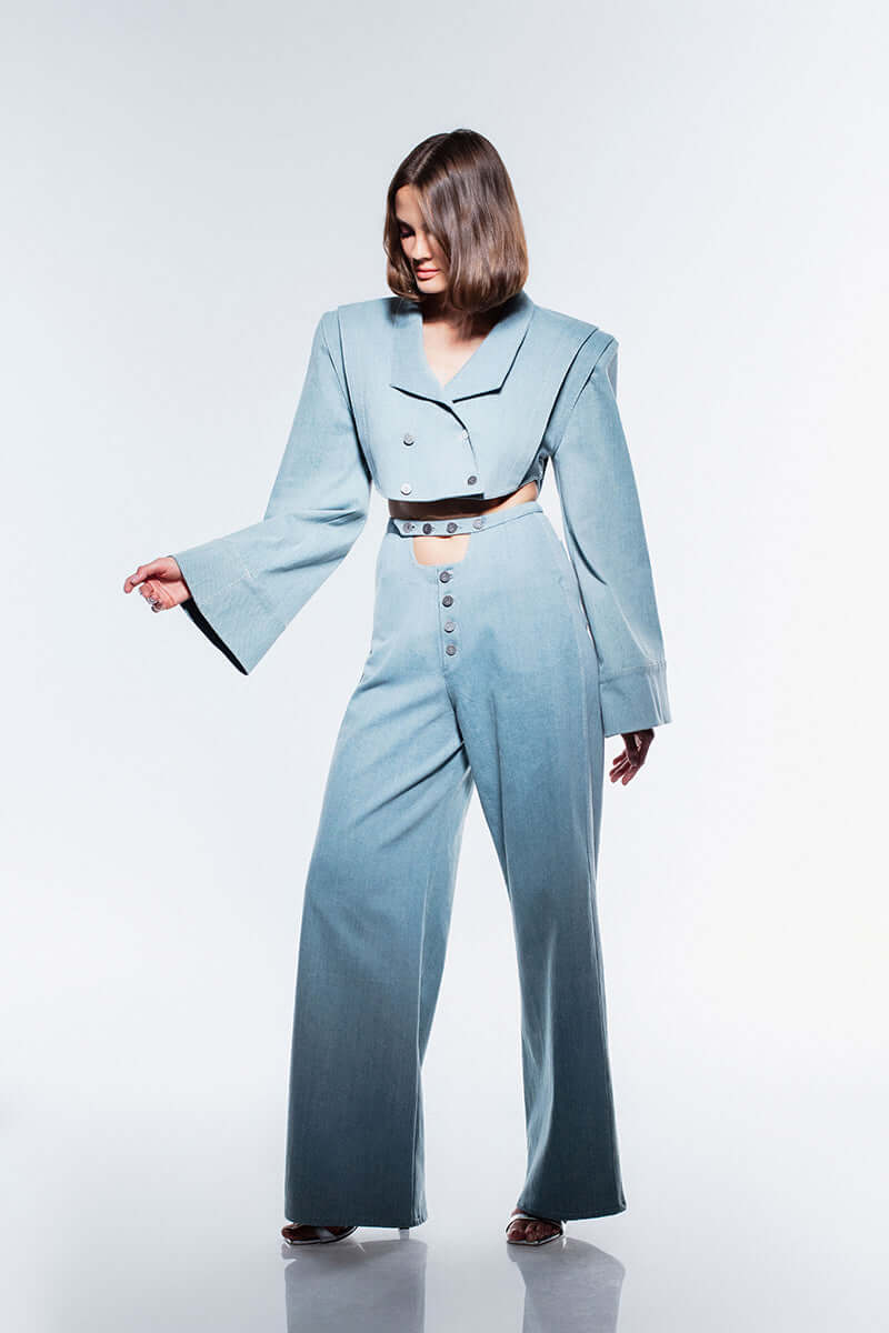 Light blue cropped denim jacket with a unique shape. It has an asymmetric button front, padded shoulders, and sleeves that flare out significantly at the wrist.
