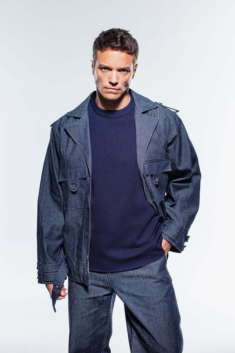 A navy blue denim jacket with white contrast stitching, featuring a front zipper, chest pockets, and a belted hem with a silver buckle.