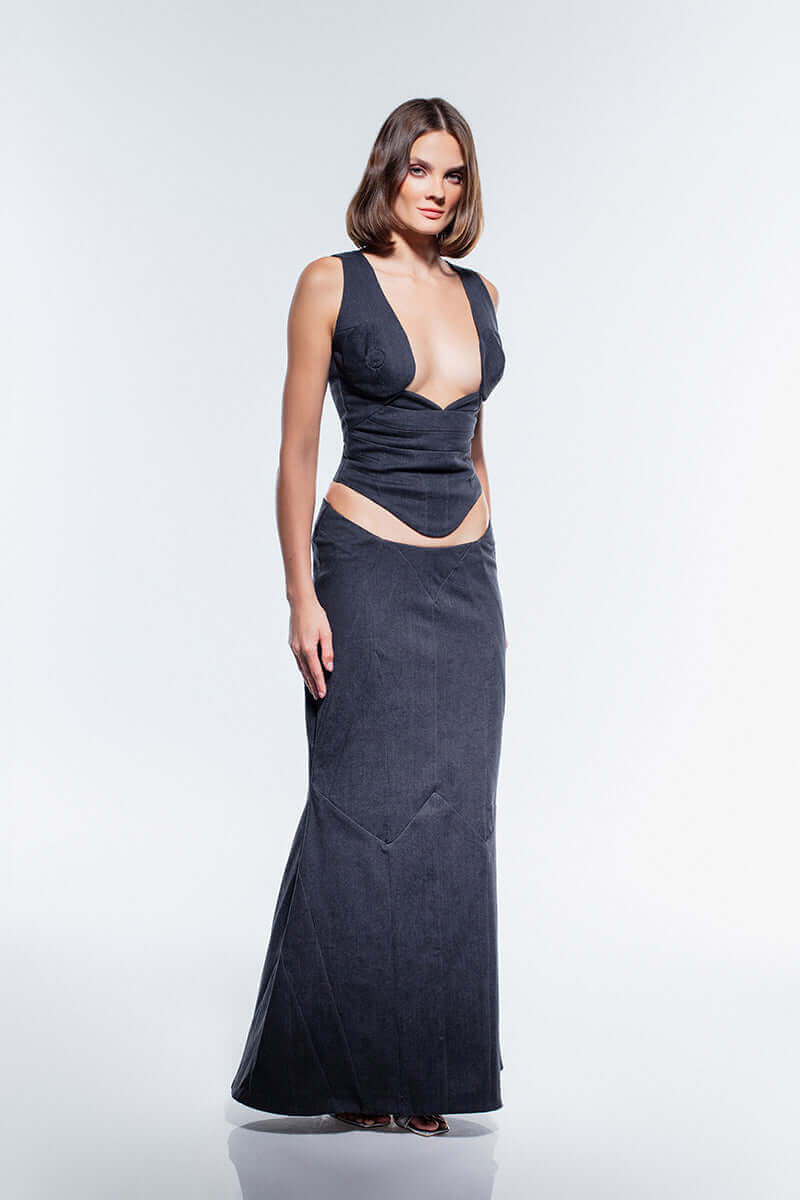 A high-waisted, dark denim maxi skirt with a subtle pinstripe pattern. The skirt has unique V-shaped seams on the front, adding to its structured design.