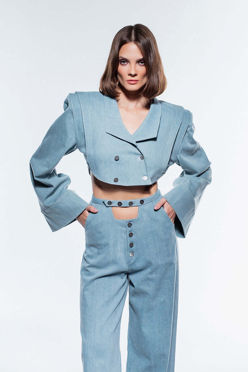 A cropped, ice blue denim jacket with wide, flared sleeves. It has an asymmetric double-breasted button closure and prominent shoulder pads.